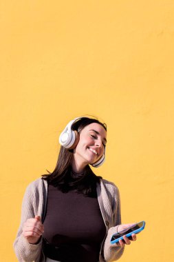 Kadın kulaklıkla telefondan müzik dinleyerek mutlu bir şekilde dans ediyor. Arka planda sarı bir duvar, ritim ve pozitif insanlar kavramı, metin için kopyalama alanı.