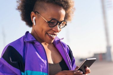 Kulaklık, teknoloji ve spor yaşam tarzıyla cep telefonuyla müzik çalan Afro-Amerikan kadın sporcunun portresi.