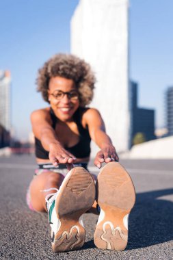 Afro-Amerikalı bir kadının, şehir parkında bacaklarını açarken çekilmiş dikey fotoğrafı, sağlık ve spor yaşam tarzı kavramı, spor ayakkabılarına seçici odaklanma ve metin için metin alanı