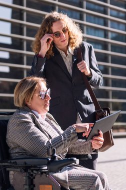 Tekerlekli sandalye kullanan kadın, finans bölgesinde iş adamına tablet gösteriyor. Çeşitlilik kavramı ve kentsel yaşam tarzı.