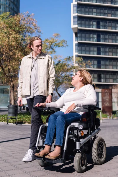 woman using electric wheelchair with a young man talking happy as they walk through the city, concept of friendship and diversity