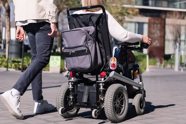 stock image cropped rear view of an unrecognizable woman using wheelchair with a man taking a walk by the city, concept of friendship and diversity