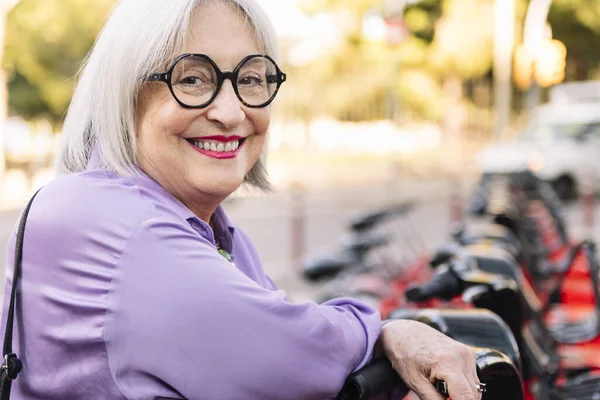 portrait of a smiling senior woman leaning on a rental bike in the parking row, concept of sustainable mobility and active lifestyle in elderly people, copy space for text