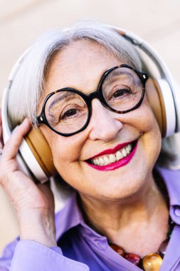 Kameraya bakan, kulaklıkla müzik dinlemekten keyif alan, yaşlı insanların boş vakitleri ve aktif bir yaşam tarzı olan gülümseyen yaşlı bir kadının portresini yakından çek.