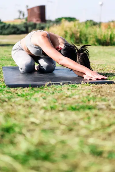 young woman in sportswear kneeling on her yoga mat doing back stretching exercises on the grass in the park, active and healthy lifestyle concept