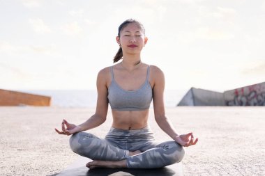 Genç Asyalı kadın deniz kenarında meditasyon yapıyor yoga minderinde oturuyor, zihinsel rahatlama ve sağlıklı yaşam tarzı kavramı.