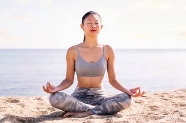 Genç Asyalı kadın plajda meditasyon yapıyor bacak bacak üstüne atmış oturuyor, zihinsel rahatlama ve sağlıklı yaşam tarzı kavramı