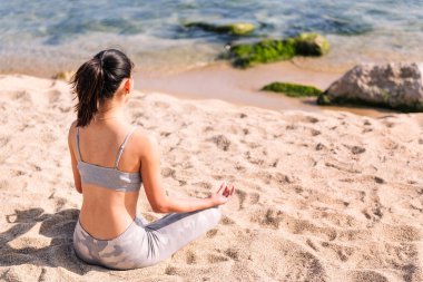 Spor kıyafetleri içinde tanınmayan genç bir kadın sahilde meditasyon yapıyor bacak bacak bacak üstüne atmış, zihinsel rahatlama ve sağlıklı yaşam tarzı kavramı ile oturuyor.
