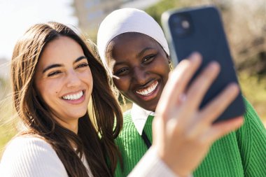 Mutlu gülümseyen ve cep telefonuyla selfie çeken iki genç bayan arkadaş, çeşitlilik kavramı ve modern yaşam tarzı.