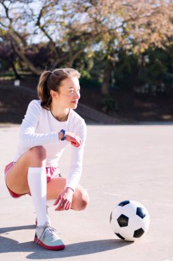 Bir futbol topunun yanında çömelmiş genç beyaz kadın bir futbol sahasında oynamaya hazır, spor anlayışı ve aktif yaşam tarzı.