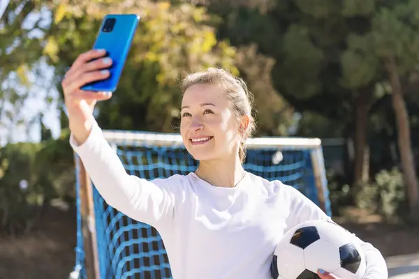 stock image smiling young caucasian woman taking a selfie photo with mobile phone while holding a soccer ball in a urban field, concept of social media and football lifestyle