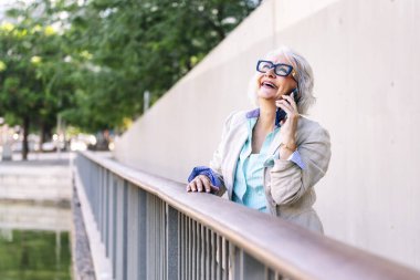 Şık, olgun bir kadın cep telefonuyla konuşurken mutlu bir şekilde gülüyor. Şehir parkında korkuluklara yaslanıyor, iletişim teknolojisi kavramı ve modern yaşam tarzı yetişkinlik çağında, metin metni için kopyalama alanı.