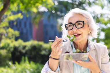Şehir parkında açık havada salata yiyen orta yaşlı bir kadının portresi. Yetişkinlikte sağlıklı yiyecek ve aktif yaşam biçimi kavramı.