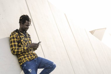 Gülümseyen genç siyah adam telefonuna mesaj yazıyor modern beton bir yapıya yaslanıyor, yaşam tarzı ve teknoloji kavramına.