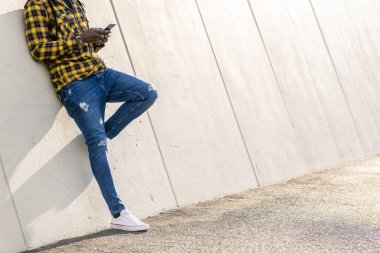 Tanımlanamayan genç siyahi adam telefonuna mesaj yazıyor modern beton bir yapıya yaslanıyor, yaşam tarzı ve teknoloji kavramına.