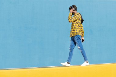 Tanımlanamayan, kulaklıklı, sarı bir duvar boyunca yürüyen, arka planda mavi bir duvar olan, elinde akıllı telefonu, teknoloji ve yaşam tarzı konseptiyle tanınmayan genç bir siyahi. Metin için kopyalama alanı.