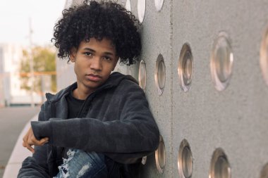 Kentsel yaşam tarzı kavramı olan şehir sokağında beton duvarın yanında oturmuş kameraya bakan kıvırcık saçlı Afrikalı Amerikalı hippi bir genç.