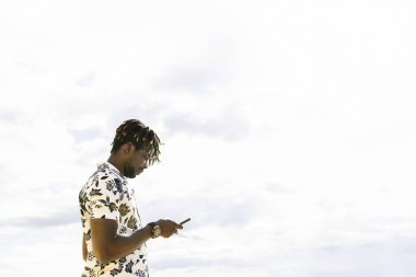 Arka planda gökyüzü, teknoloji ve yaşam tarzı konsepti olan cep telefonuna bakan şık genç siyahi adam, Tex için kopyalama alanı