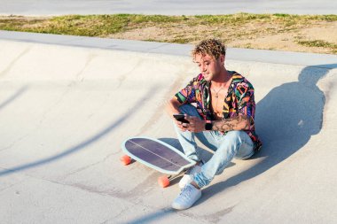 Milenyum kıvırcık saçlı, renkli gömlekli, kot pantolonlu kaykaylı, beton rampada oturan kaykaylı ve cep telefonlu yakışıklı adam.