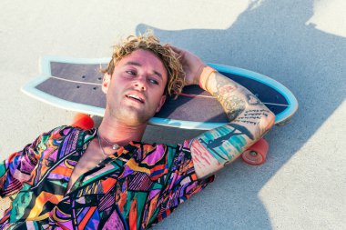 Havalı renkli tişörtlü genç kaykaycı kaykay pistinde dinleniyor.