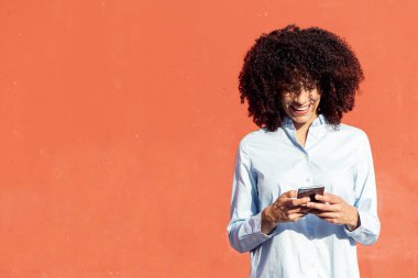 Turuncu arka planda dururken akıllı telefondan mesaj atan resmi tişörtlü etnik kadın girişimci
