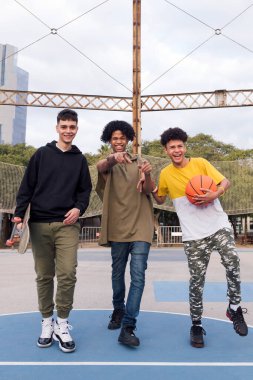 Basketbol ve kaykaylı kendinden emin üç genç erkek parkta, gençlik ve şehir yaşam tarzında bir spor sahasında eğlenirken şakalaşıp gülüyorlar.