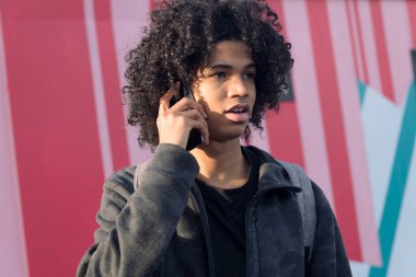 Renkli duvarın önünde cep telefonuyla konuşan kıvırcık saçlı Afro-Amerikan ergenin portresi, iletişim teknolojisi ve genç yaşam tarzı kavramı