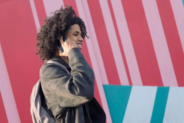 Gülümseyen kıvırcık saçlı Afrikalı Amerikalı genç cep telefonuyla konuşuyor renkli duvarların önünde yürüyor, iletişim teknolojisi kavramı ve genç yaşam tarzı