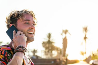 Milenyum güzeli kıvırcık saçlı, şık kıyafetli, cep telefonuyla konuşan yaz günü açık hava parkında duran adamın yan görüntüsü.