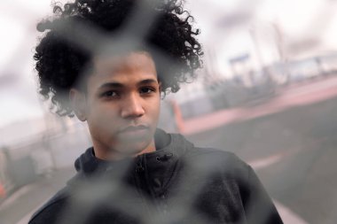 Düşünceli Afro-Amerikan erkek portresi. Kıvırcık saçlı, kameraya bakan şehir sokaklarında metal tel örgülerin arkasında duran ergenlik ve akıl sağlığı kavramı.
