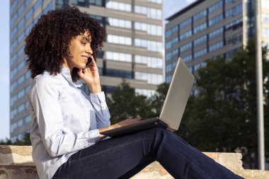 Parktaki taş bankta oturan ve akıllı telefonla konuşurken uzaktan çalışan ve dizüstü bilgisayar kullanan mutlu kadın girişimcinin yan görüntüsü