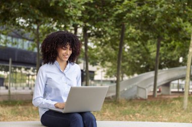 Parktaki taş bankta oturan ve dizüstü bilgisayarla uzaktan çalışan kıvırcık saçlı mutlu kadın girişimci.