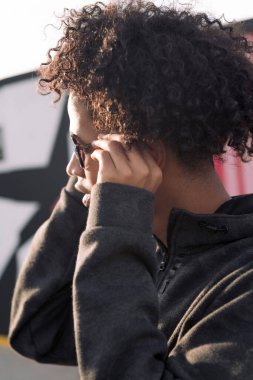 Kendine güvenen kıvırcık saçlı Afro-Amerikan genç erkek güneş gözlüklerini düzeltirken renkli şehir geçmişine karşı duruyor, genç yaşam tarzı kavramı