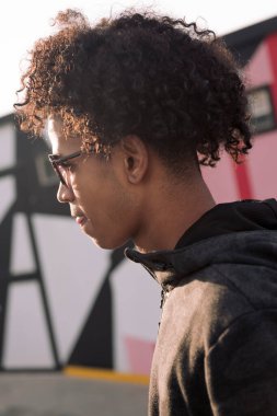 Kendine güvenen kıvırcık saçlı, modaya uygun güneş gözlüklü Afro-Amerikan bir gencin renkli şehir geçmişinin önünde yürürken çekilmiş portresi.