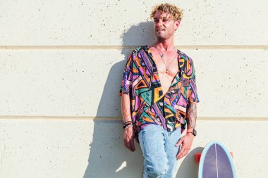 Kaygısız erkek kaykaycı modaya uygun giysiler içinde binaya yaslanıyor ve güneşli bir günde kaykayla ayakta dikiliyor.