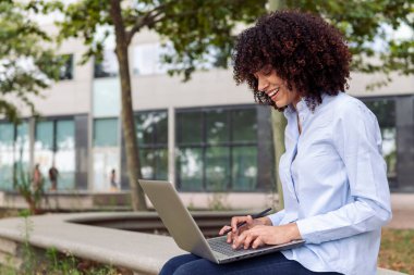 Parktaki taş bankta oturan ve dizüstü bilgisayarla uzaktan çalışan kıvırcık saçlı mutlu kadın girişimci.