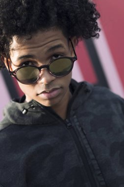 Kendine güvenen kıvırcık saçlı Afro-Amerikan genç erkek güneş gözlüklerine bakarken bulanık kentsel arka planda kameraya bakıyor.