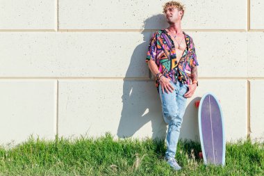 Tasasız erkek patenci, modaya uygun giysiler içinde parkta duvara yaslanıyor ve güneşli bir günde kaykayla dikiliyor.