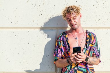 Erkek neşeli hipster güneşli bir günde duvara yaslanıyor ve sosyal medyada cep telefonuyla sohbet ediyor.