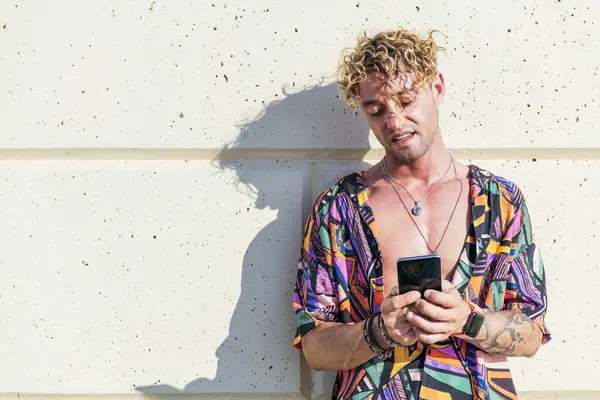 Erkek neşeli hipster güneşli bir günde duvara yaslanıyor ve sosyal medyada cep telefonuyla sohbet ediyor.