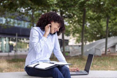 Gülümseyen iş kadını parkta taş bankta oturuyor ve bilgisayarla uzaktan çalışıyor ve akıllı bir telefonla konuşuyor.