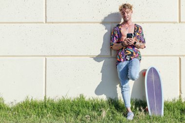 Erkek neşeli hipster parkta kaykayın yanında duvara yaslanıyor ve güneşli bir günde sosyal medyada cep telefonuyla sohbet ediyor.