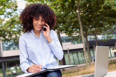gülümseyen bayan girişimci parkta taş bankında oturuyor ve akıllı telefonla konuşurken uzaktan çalışıyor ve belgelere not alıyor.