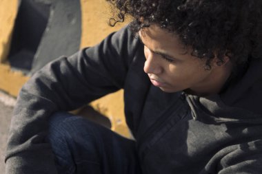 Kentsel caddede çizgili duvarın yanında oturan ciddi bir Afro-Amerikan erkek gencin portresi, ergenlik ve akıl sağlığı kavramı.
