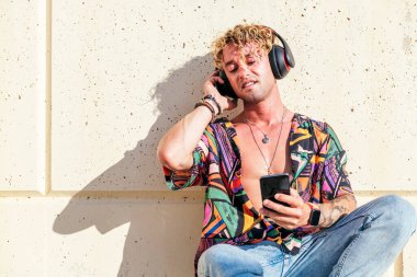 Erkek hippi sokakta oturuyor ve kapalı gözlerle kulaklıkla şarkıların tadını çıkarıyor.