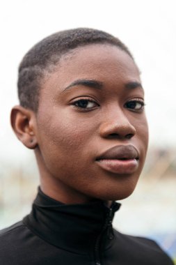 Genç, narin, kısa saçlı, arka planda bulanık kameraya bakan bir kadının portresi.