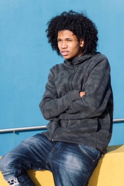 Sarı bir duvarda oturan kıvırcık saçlı, mavi arka planda kollarını kavuşturmuş, ergenlik kavramı ve genç yaşam tarzı olan Afrikalı Amerikalı ciddi bir genç.