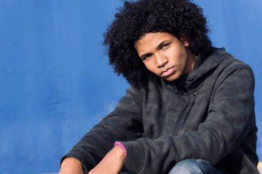 Düşünceli kıvırcık saçlı, gayri resmi giyinmiş genç siyah erkek sokakta mavi duvara yaslanırken kameraya bakıyor, spor anlayışı ve genç yaşam tarzı, metin için kopyalama alanı