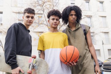 Basketbol ve kaykaylı üç farklı genç arkadaş birlikte kameraya, gençlik ve şehir yaşam tarzına bakıyorlar.