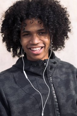 Kıvırcık saçlı Afro-Amerikan ergen bir çocuğun portresi mutlu bir şekilde gülümsüyor kulaklıklarla müzik dinliyor ve kameraya bakıyor, ergenlik kavramına ve genç yaşam tarzına bakıyor.